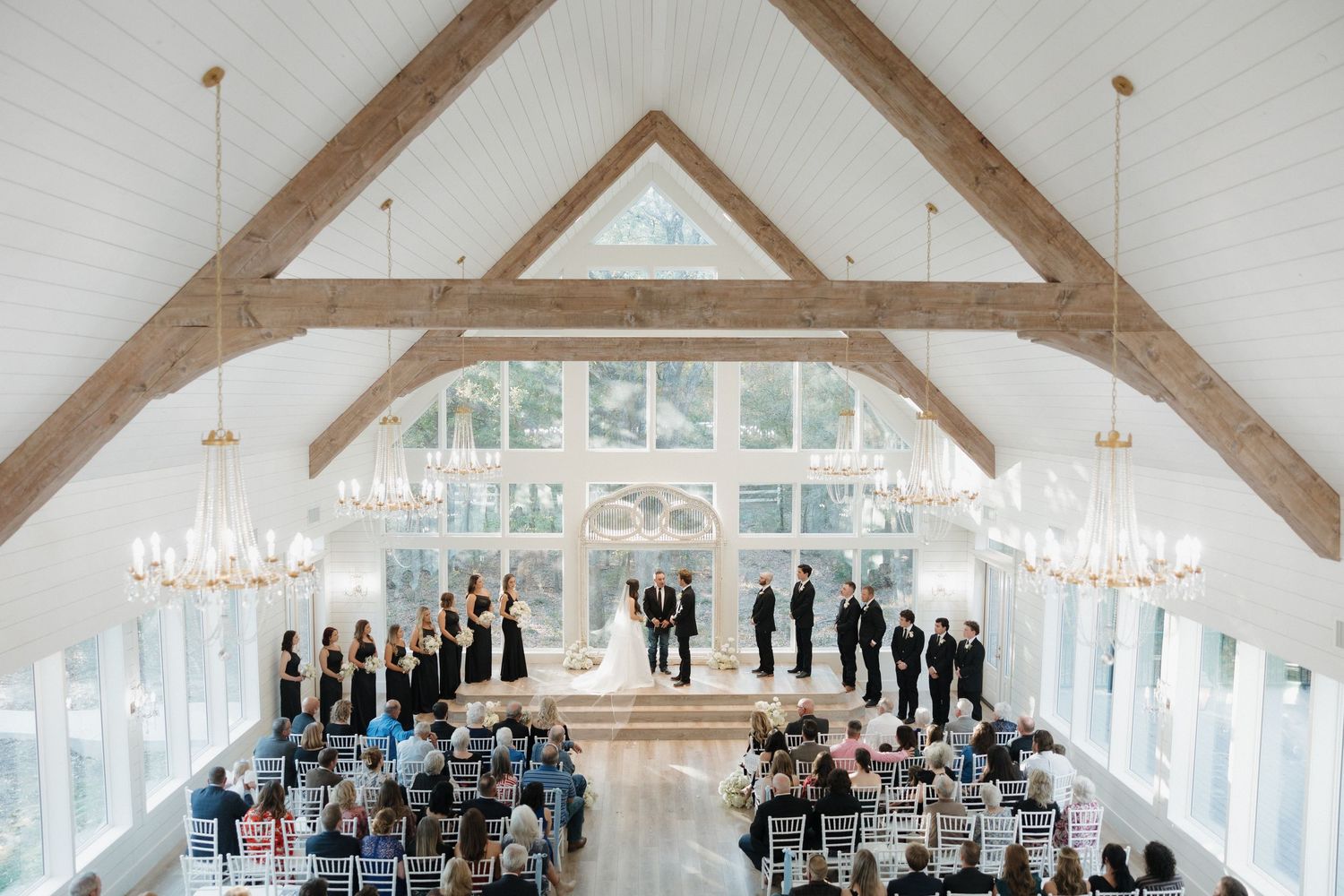 Panoramic view of an elegant white wedding chapel interior with wooden beams and crystal chandeliers during a ceremony.