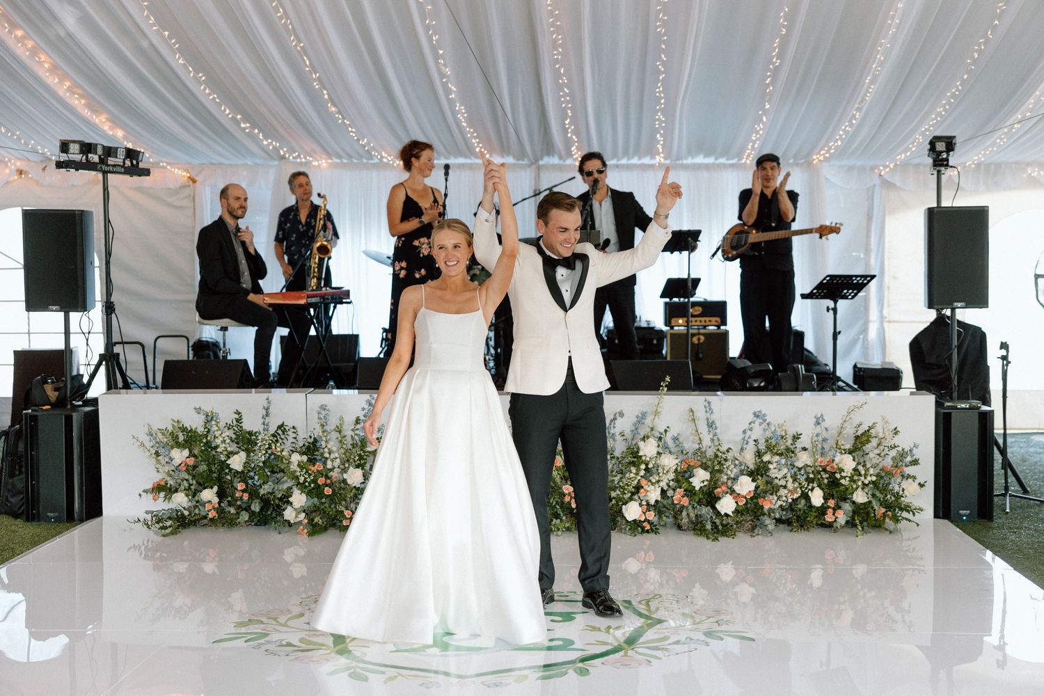 Wedding guests dancing and celebrating on a dance floor under a white tent during an outdoor reception celebration.