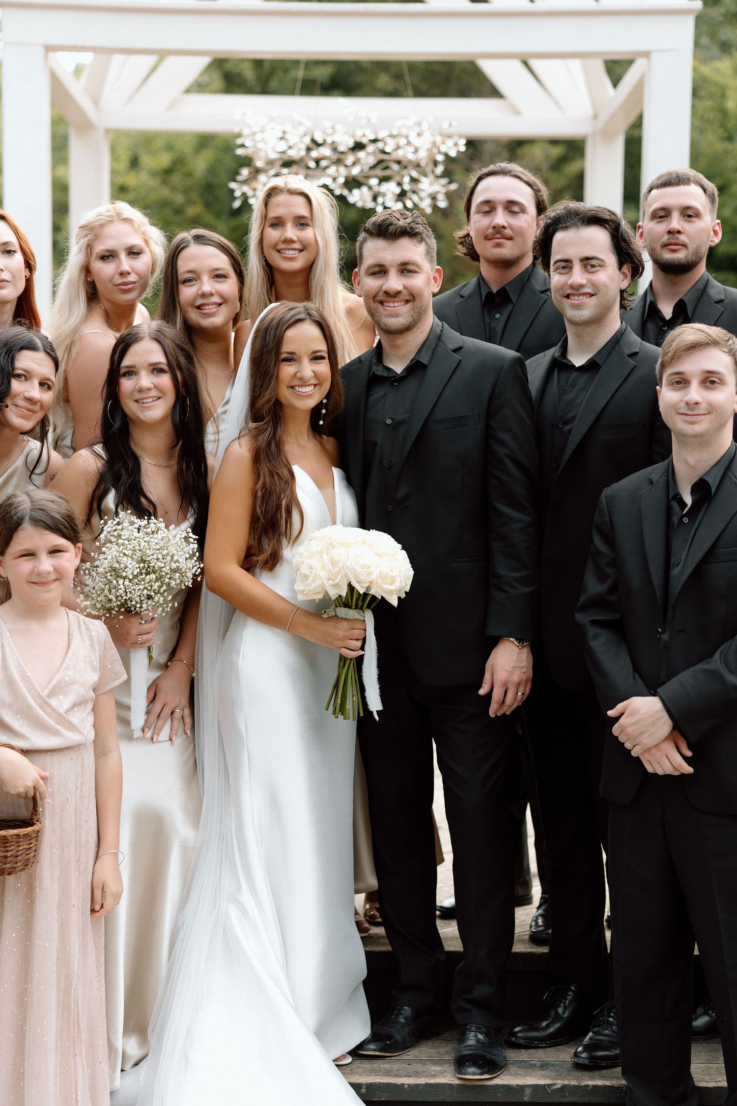 Wedding party members in formal black attire and light pink dresses pose together outdoors under a white arbor.