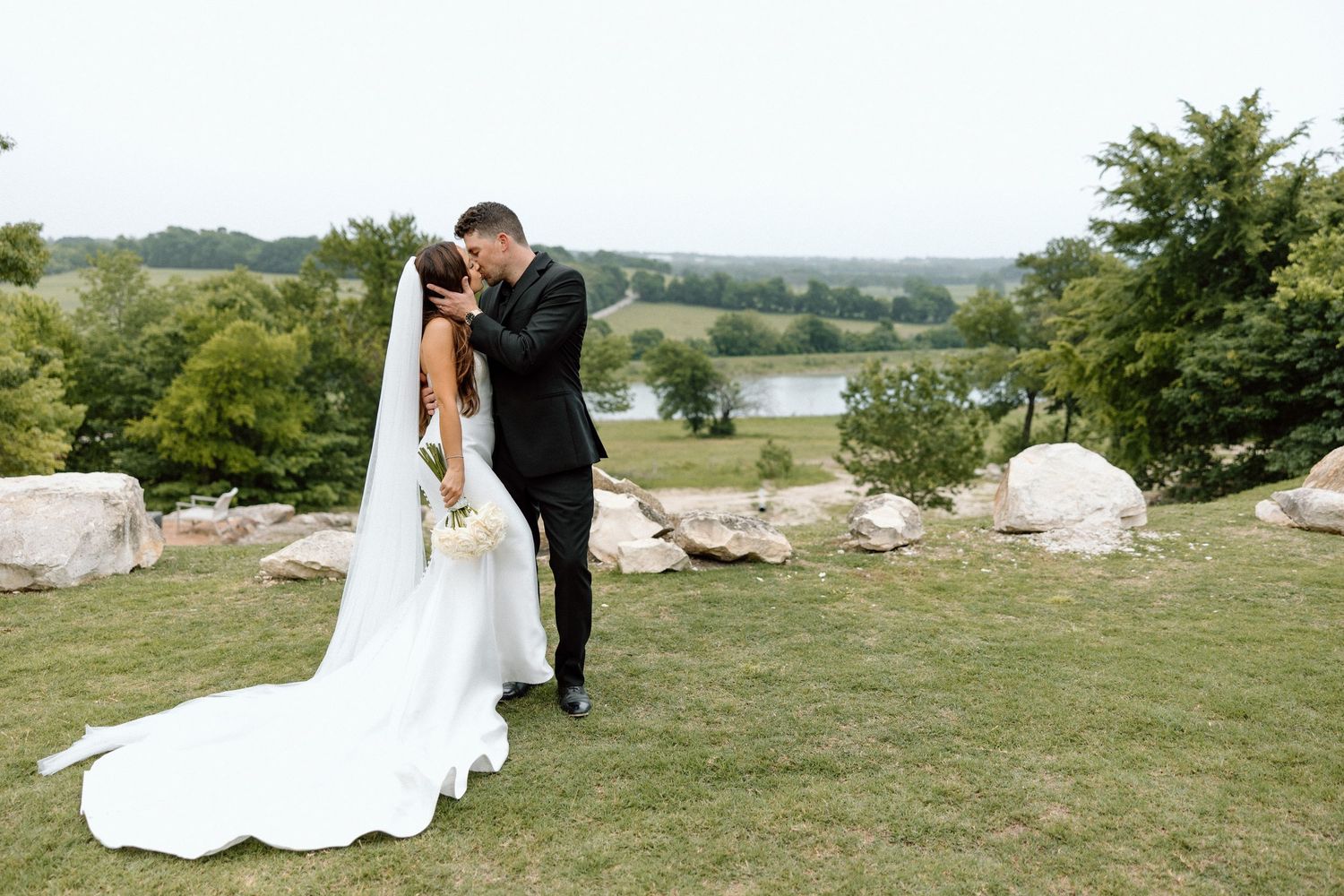 A couple shares a romantic moment on a grassy hilltop overlooking a lake, surrounded by rocks and trees.
