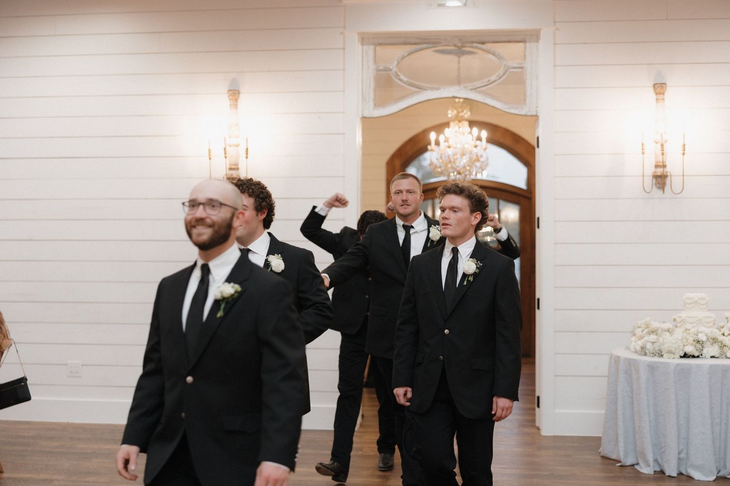 Groomsmen in black suits and ties celebrate at an elegant wedding reception venue with crystal chandeliers and wall sconces.