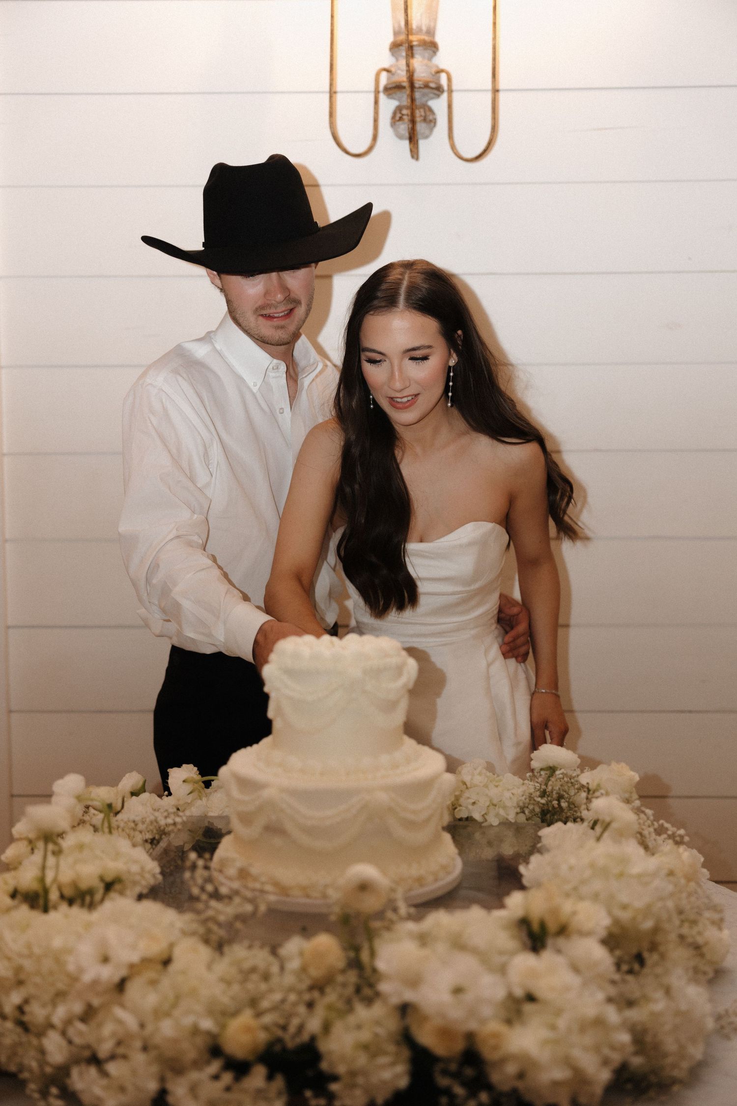 A couple cuts their white wedding cake together surrounded by delicate white floral decorations at their reception.