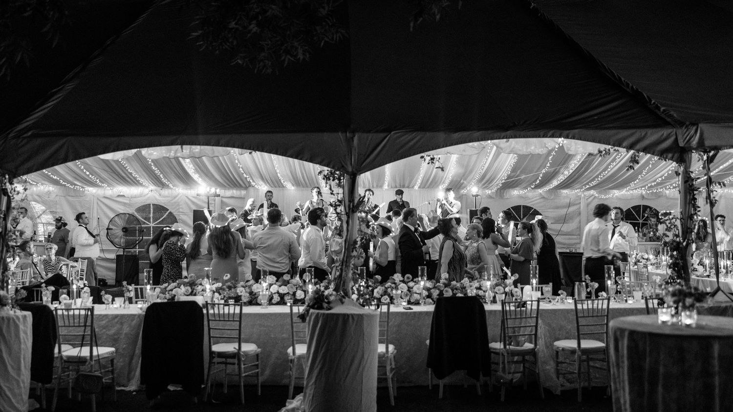 Black and white photo of a wedding reception head table under a tent with string lights and floral centerpieces.