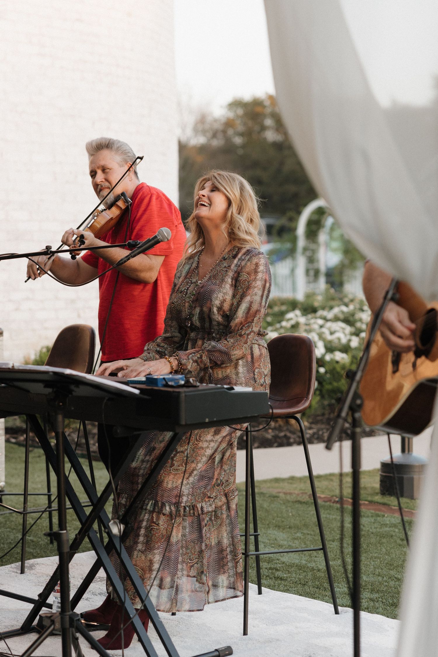 Musicians perform on keyboard and guitar at an outdoor venue with white curtains and greenery in the background.
