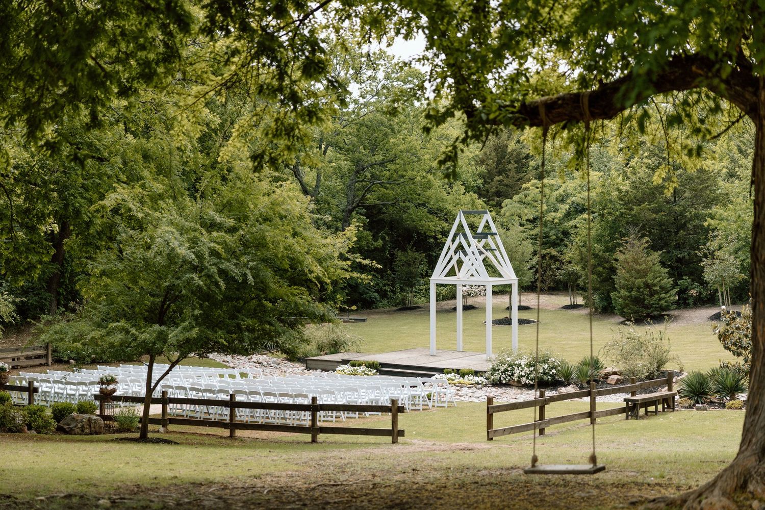 A white chapel-style wedding arch stands on a platform surrounded by wooden fences and lush green trees in a park setting.