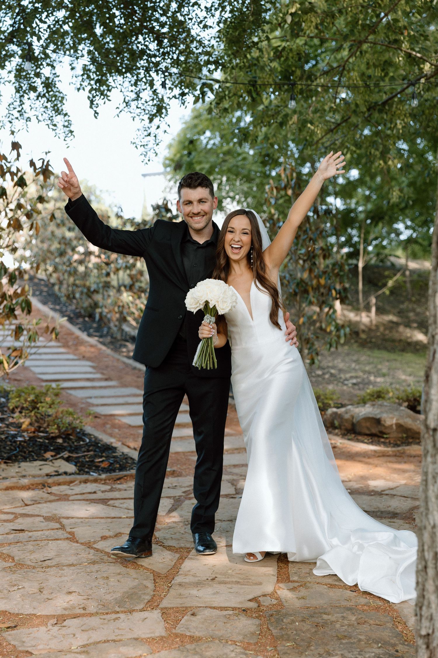 A joyful wedding couple poses playfully on stone steps outdoors surrounded by green trees.