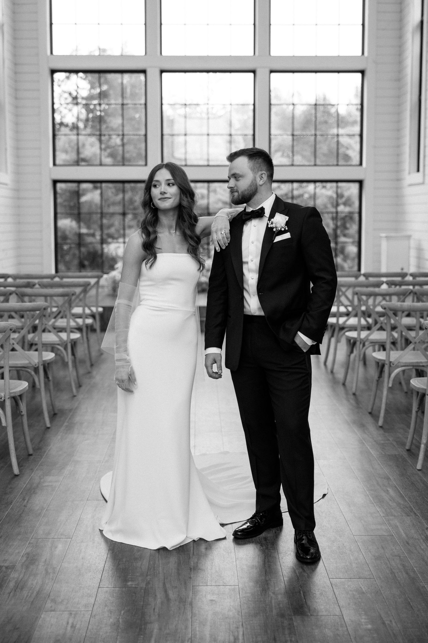 A bride and groom stand together in an elegant chapel with wooden floors and rows of chairs in black and white.