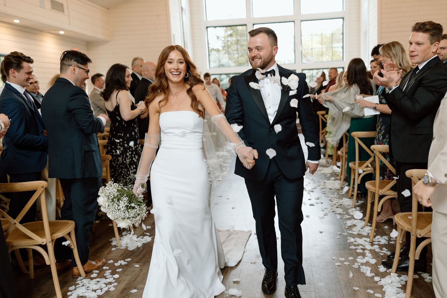 Wedding ceremony guests stand in a bright venue with large windows and wooden chairs as flower petals line the aisle.