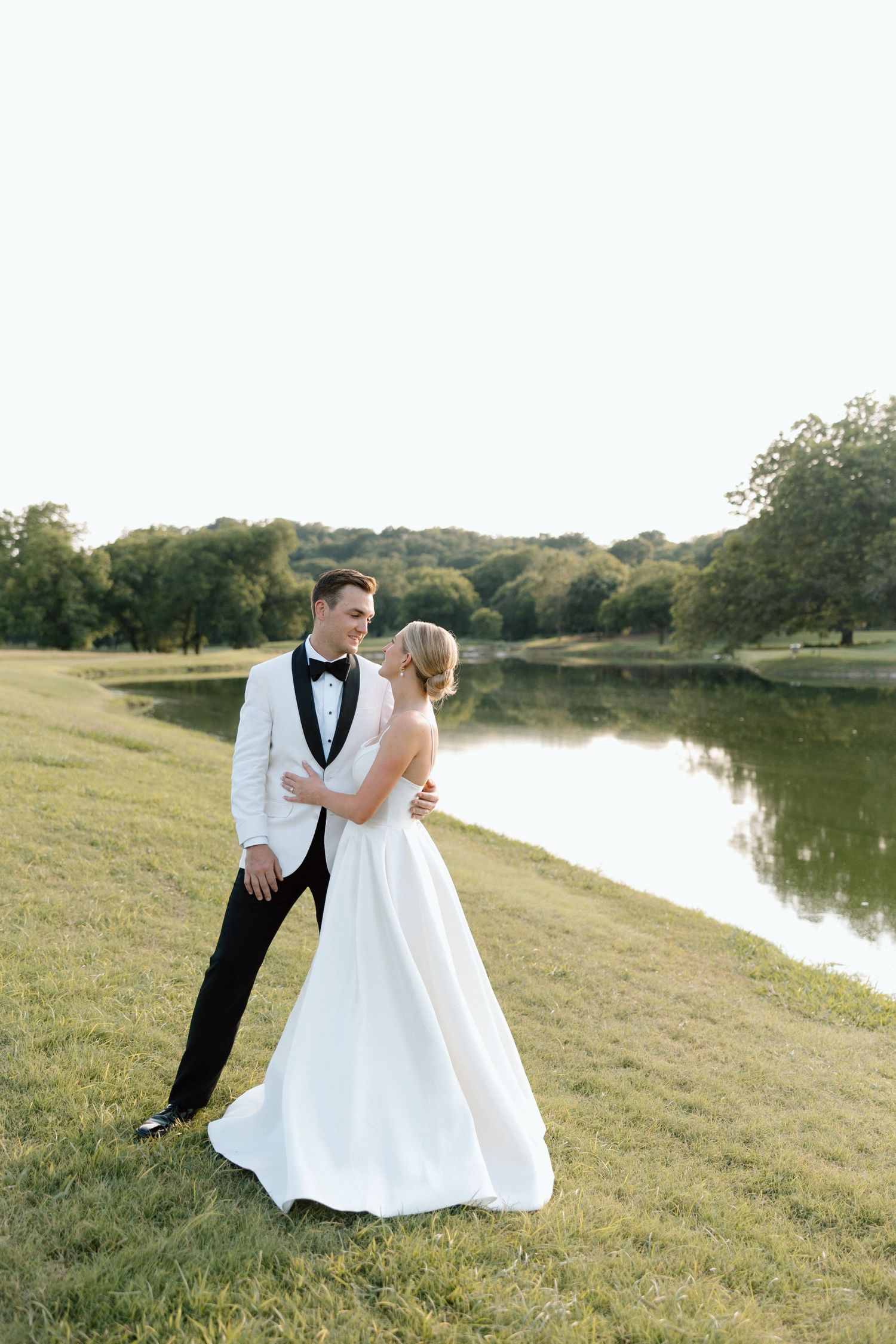 Bride and groom dance together on a grassy hillside overlooking a serene lake during their wedding photoshoot.