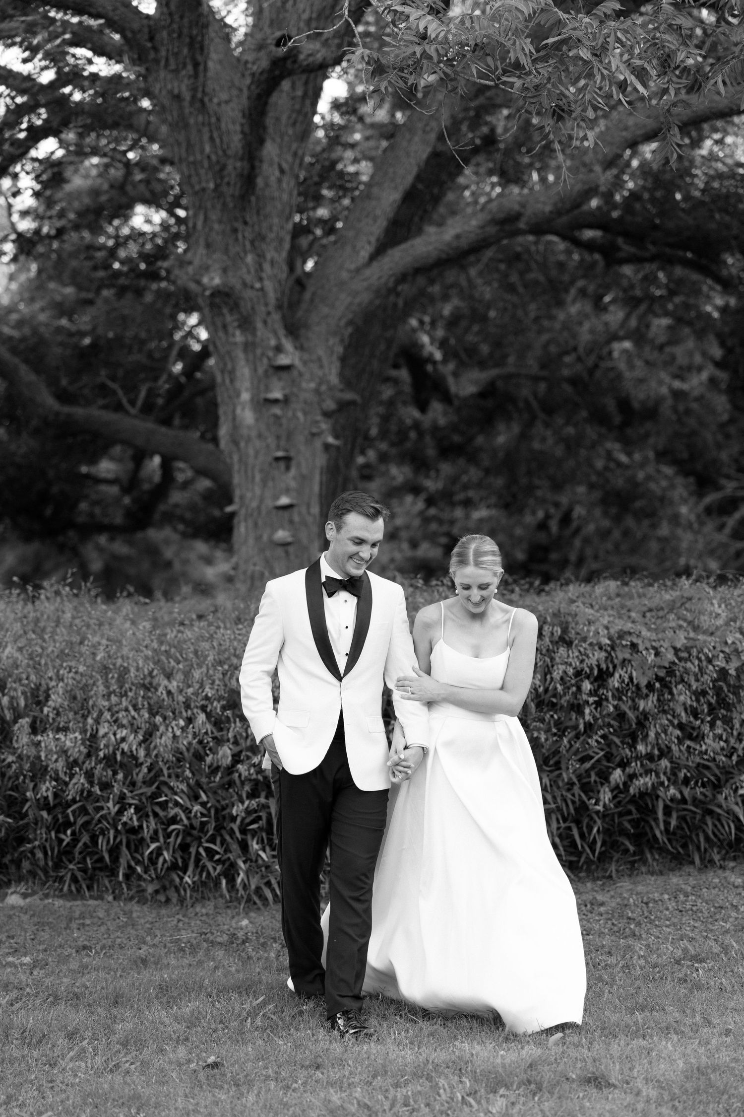 A couple in formal wedding attire walks together through a garden setting in black and white photography.