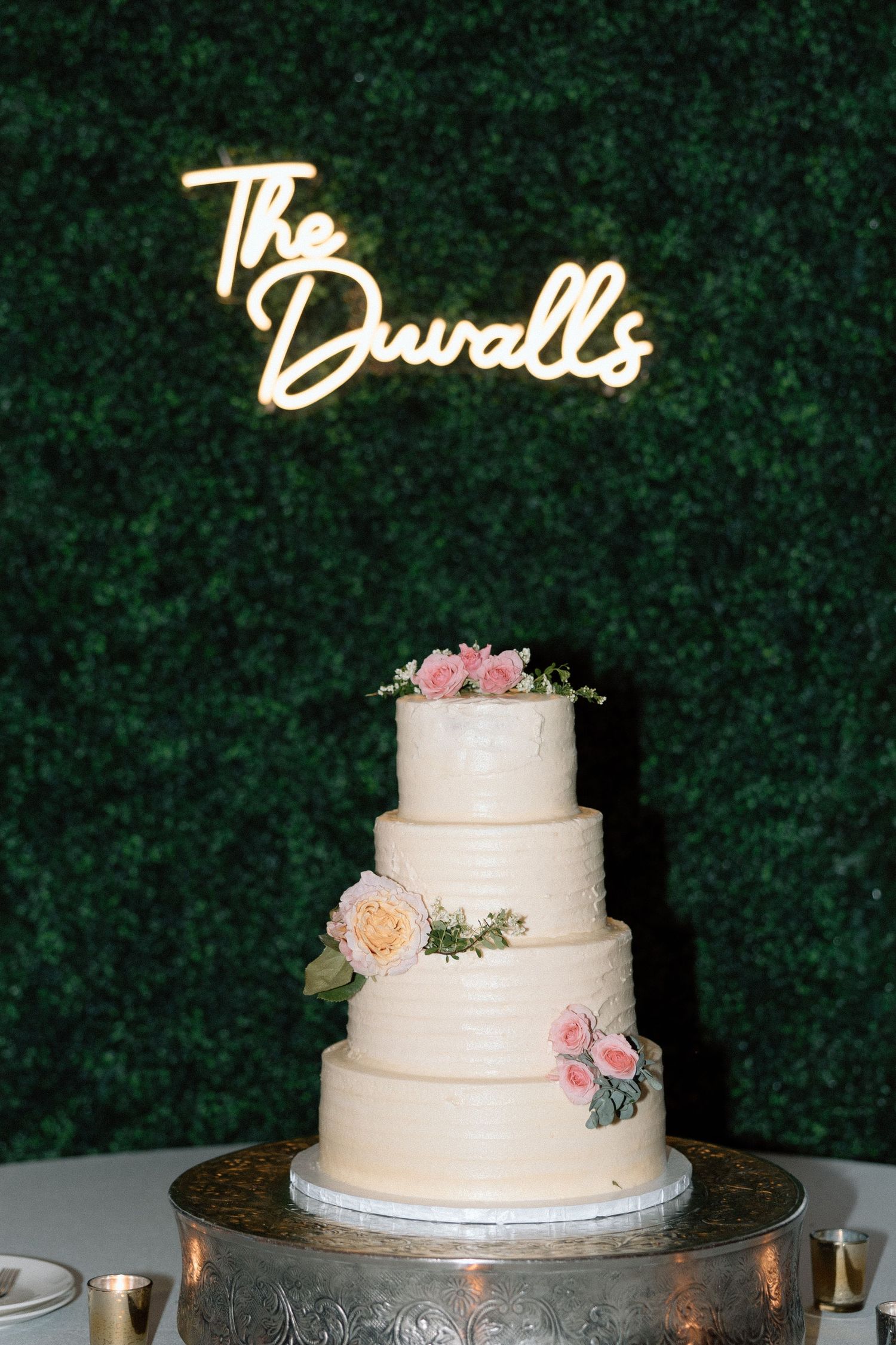 Elegant four-tier white wedding cake with pink floral accents sits beneath illuminated 'The Duvalls' sign against green backdrop.