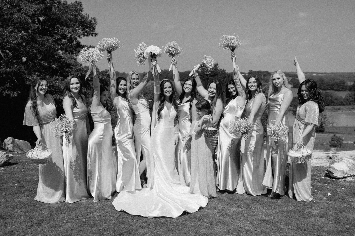 A black and white wedding photo showing a bridal party in light-colored dresses holding bouquets outdoors.