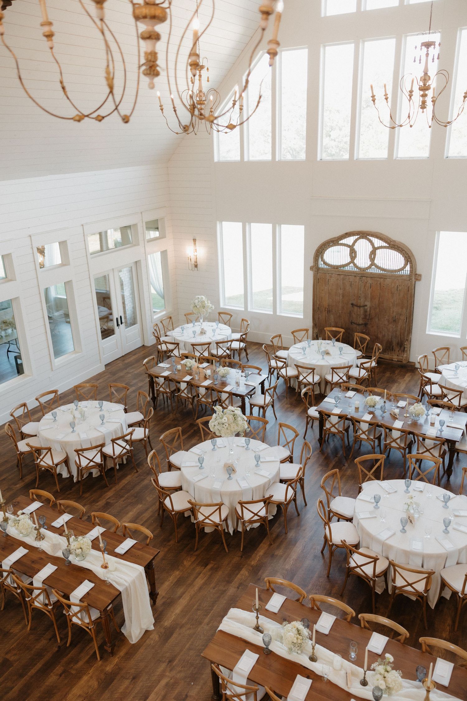Elegant wedding reception venue with round tables, white linens, wooden chairs, and chandeliers in a bright white room.