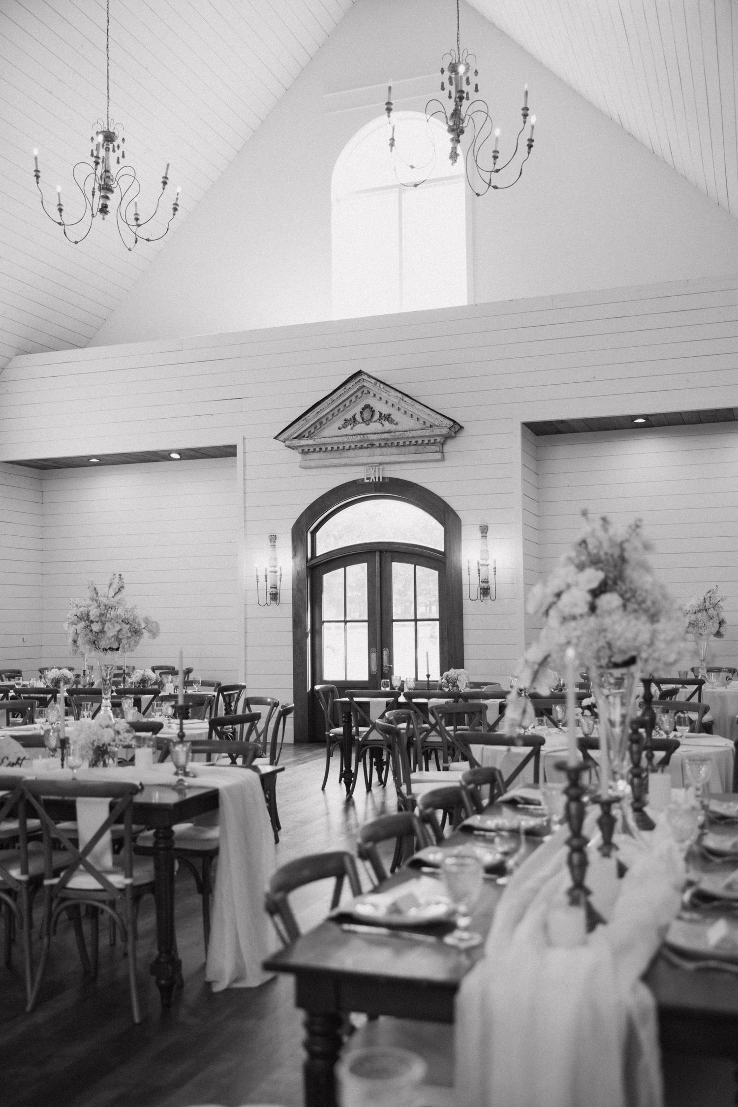 Black and white panoramic view of an elegant wedding reception venue with chandeliers and table settings in a vaulted room.