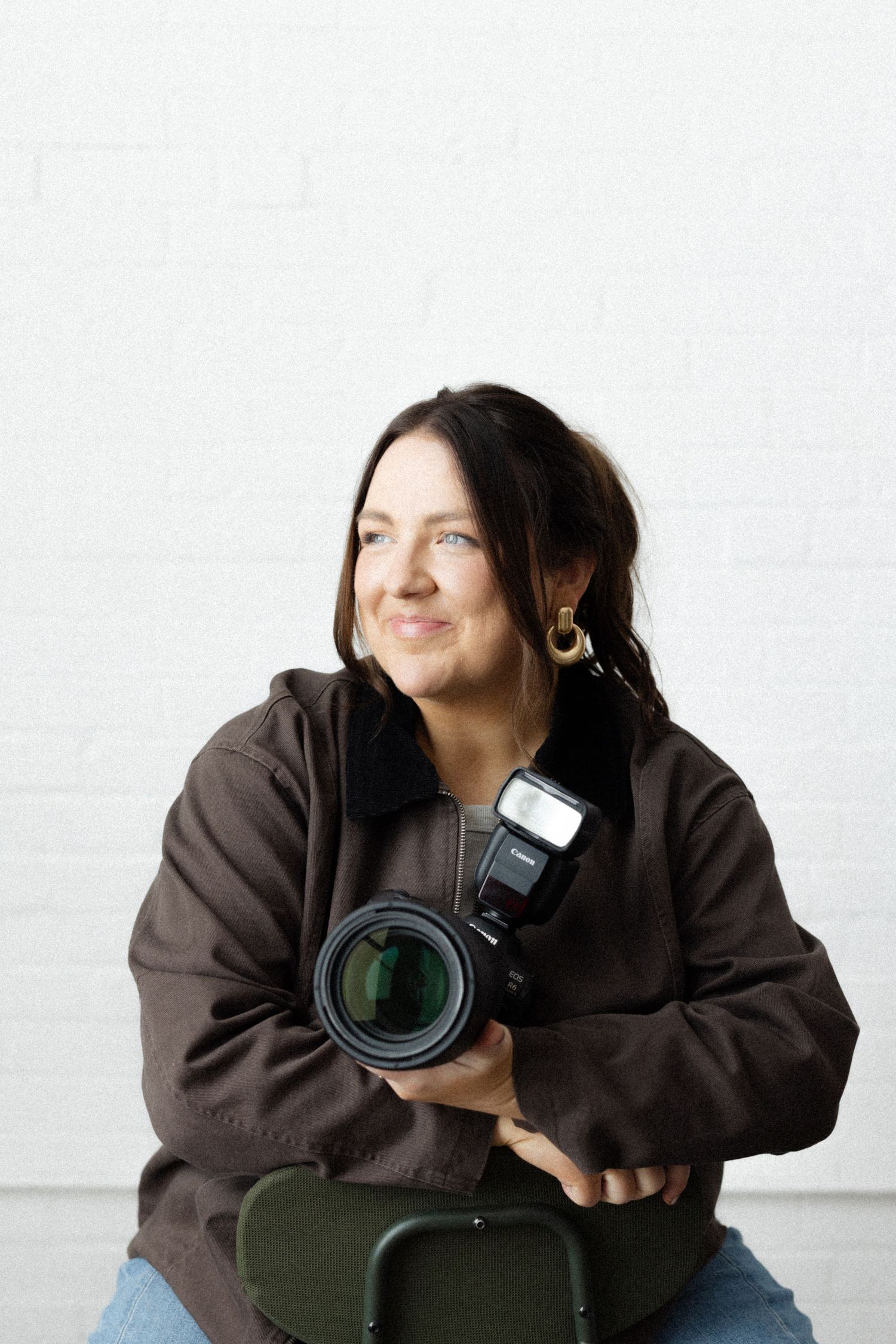 A smiling female photographer holding a DSLR camera with flash, seated against a white brick wall.