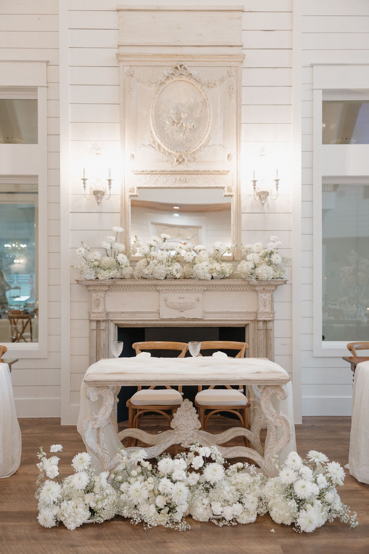 Elegant white fireplace mantel with ornate details and white floral arrangements in a luxurious wedding venue setting.