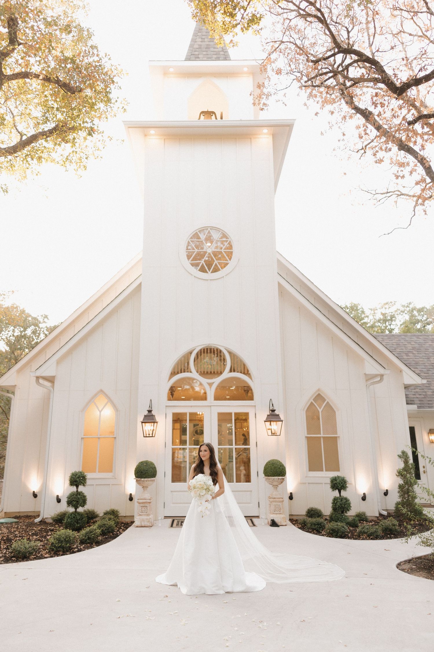 Elegant white chapel with Gothic windows and round stained glass features surrounded by autumn trees at sunset.