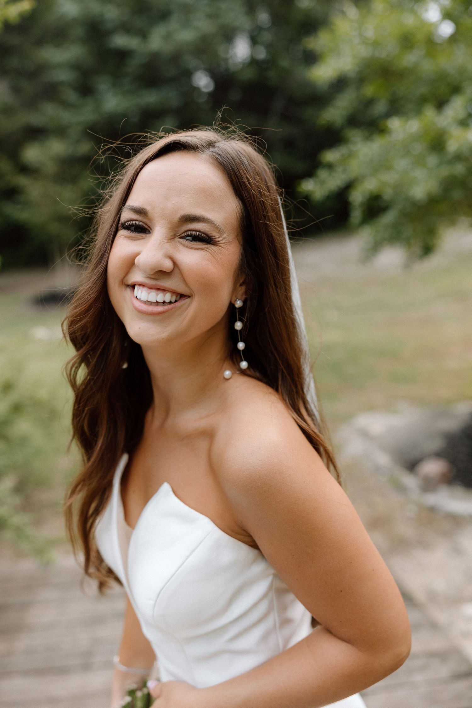 A person in a white formal dress poses outdoors against a natural background with trees and greenery.