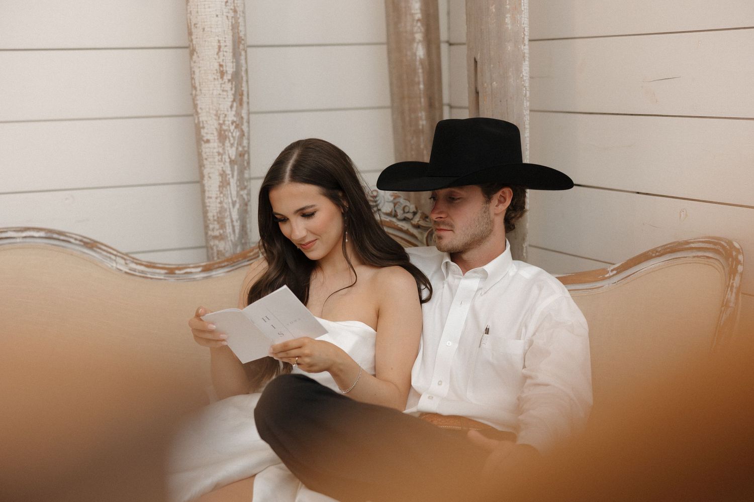 A couple in western-inspired attire sits together on a vintage couch while reading papers.