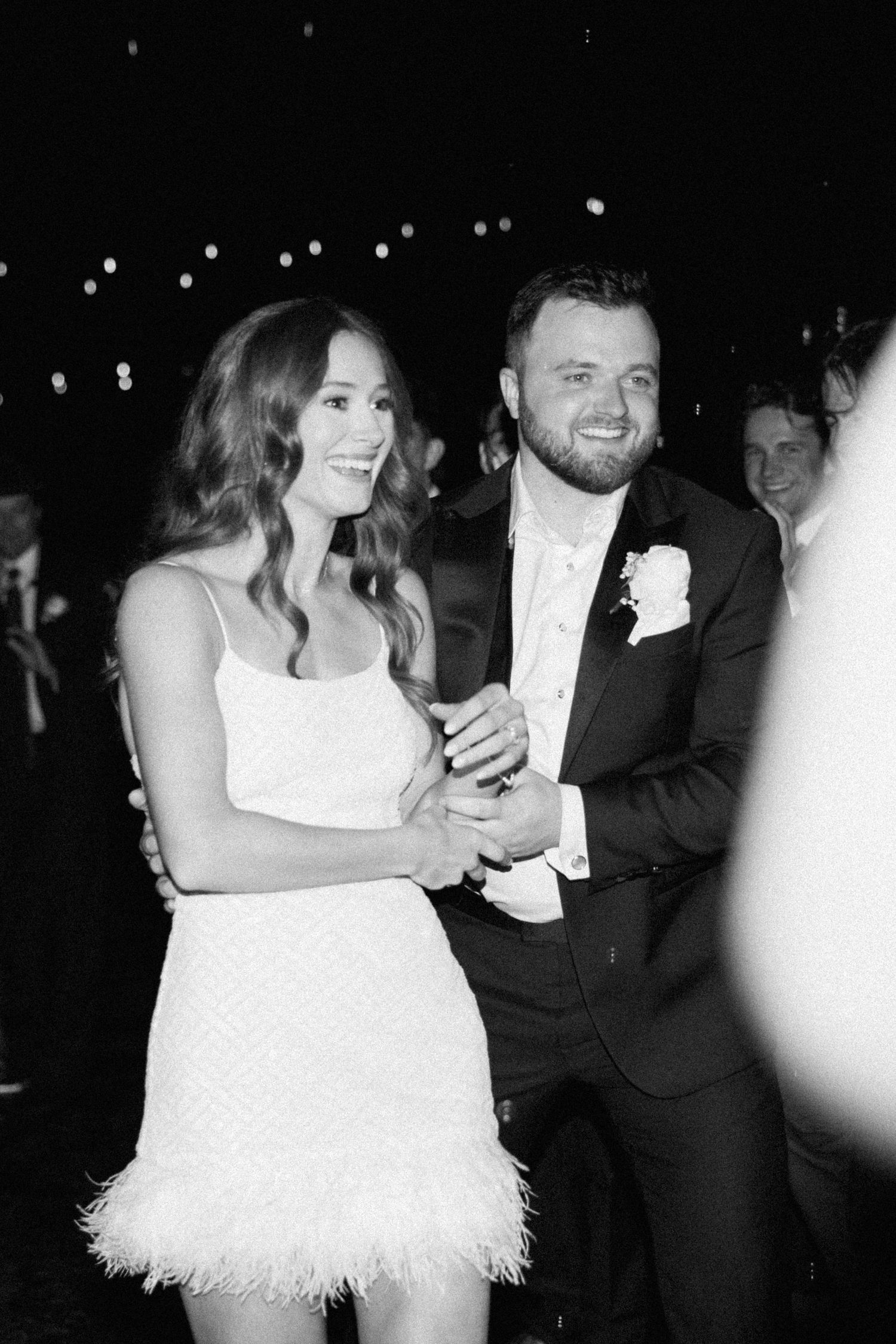 A couple dances together at a wedding reception, with the bride wearing a short white feathered dress.