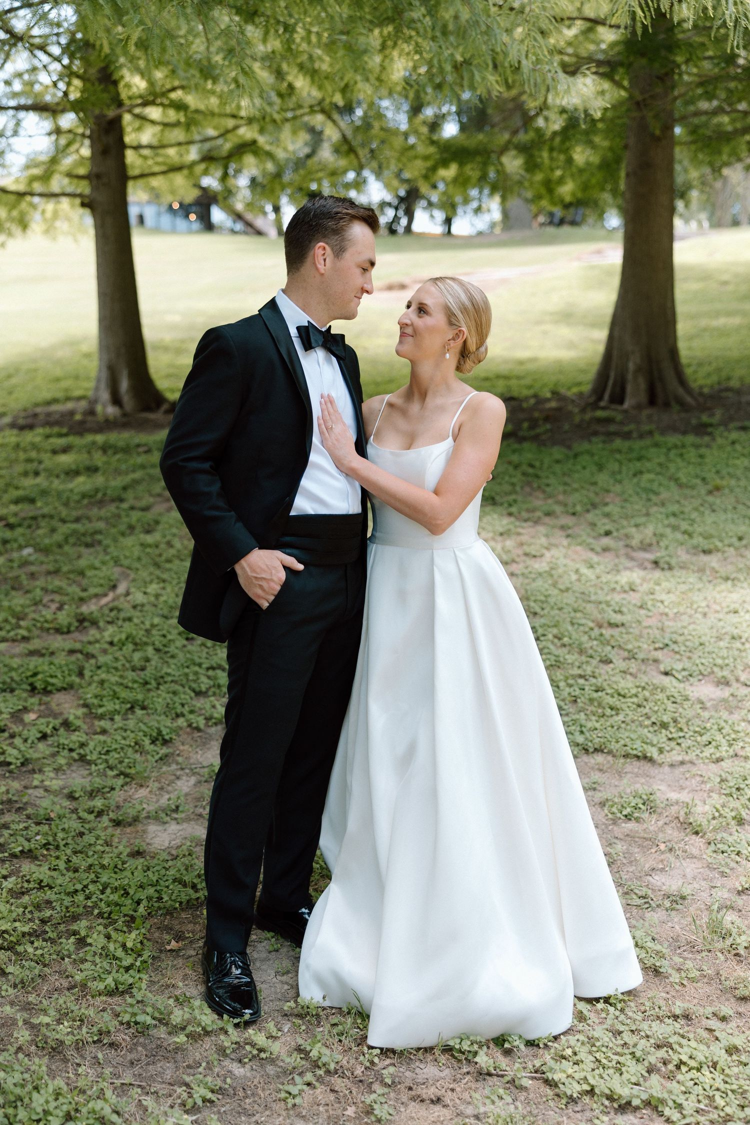 A couple in wedding attire shares an intimate moment on a grassy hill under tall trees.