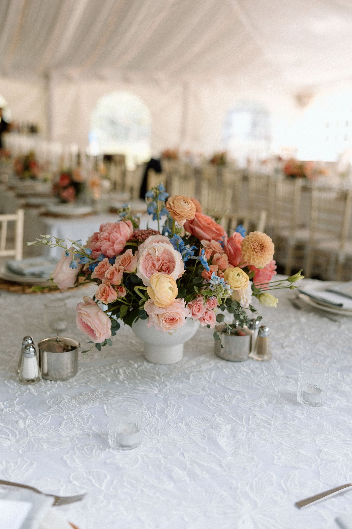 Elegant wedding table centerpiece with peach and pink roses in white vase on lace tablecloth under white tent.