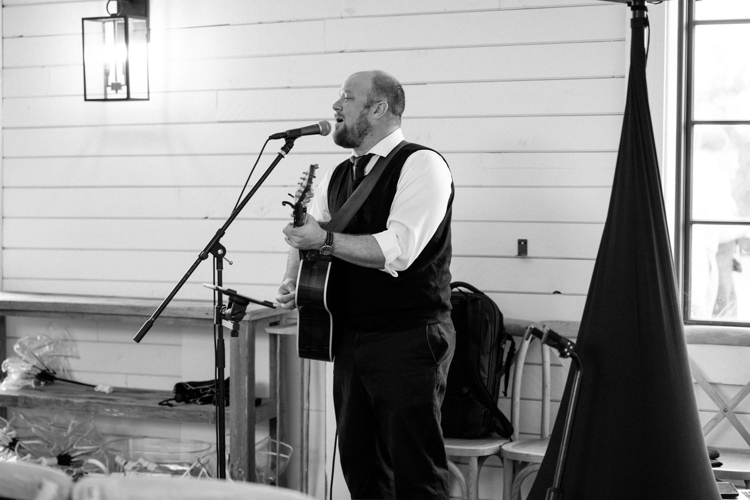 Musician performs on stage in a white-walled venue with microphone and black backdrop.