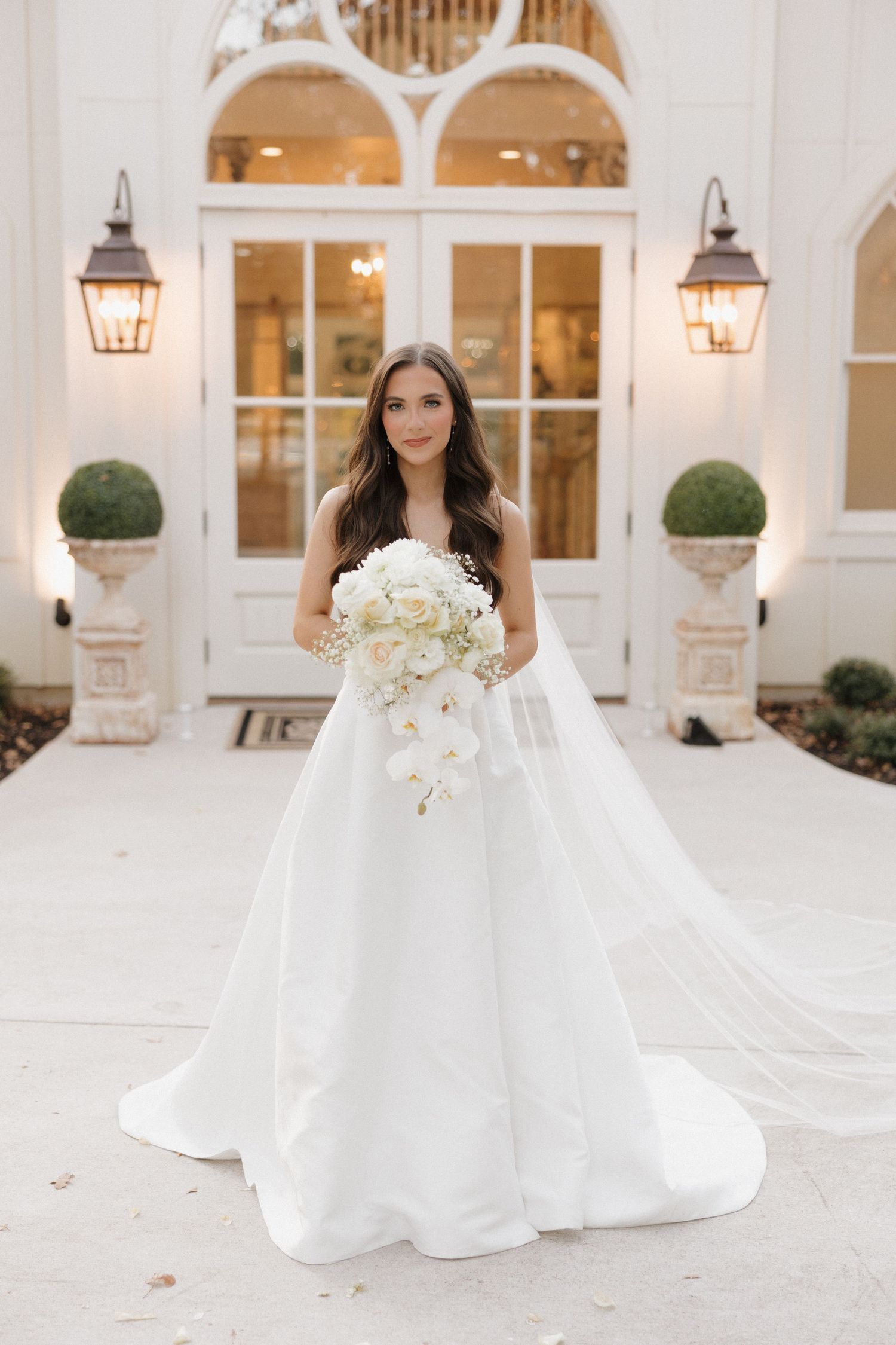 A bride in a classic white ballgown poses at an elegant venue with arched windows and lantern lighting.