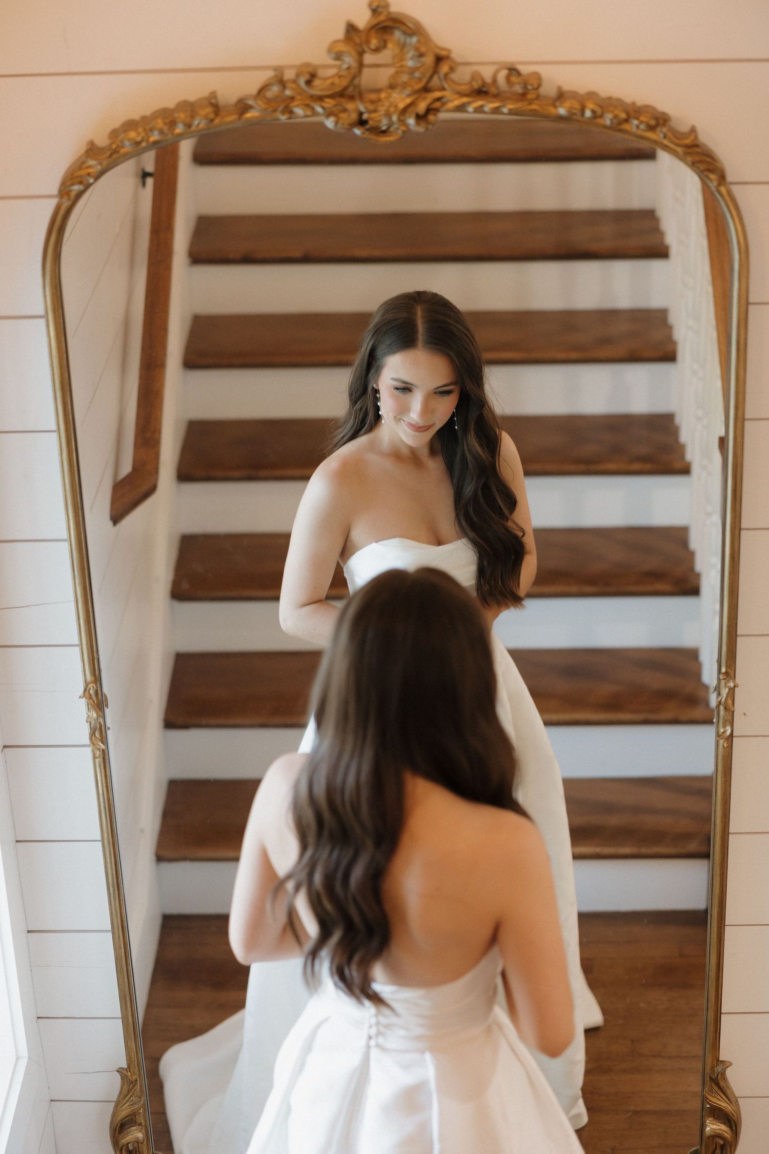 Reflection in ornate gold mirror shows person in white dress standing on wooden staircase.