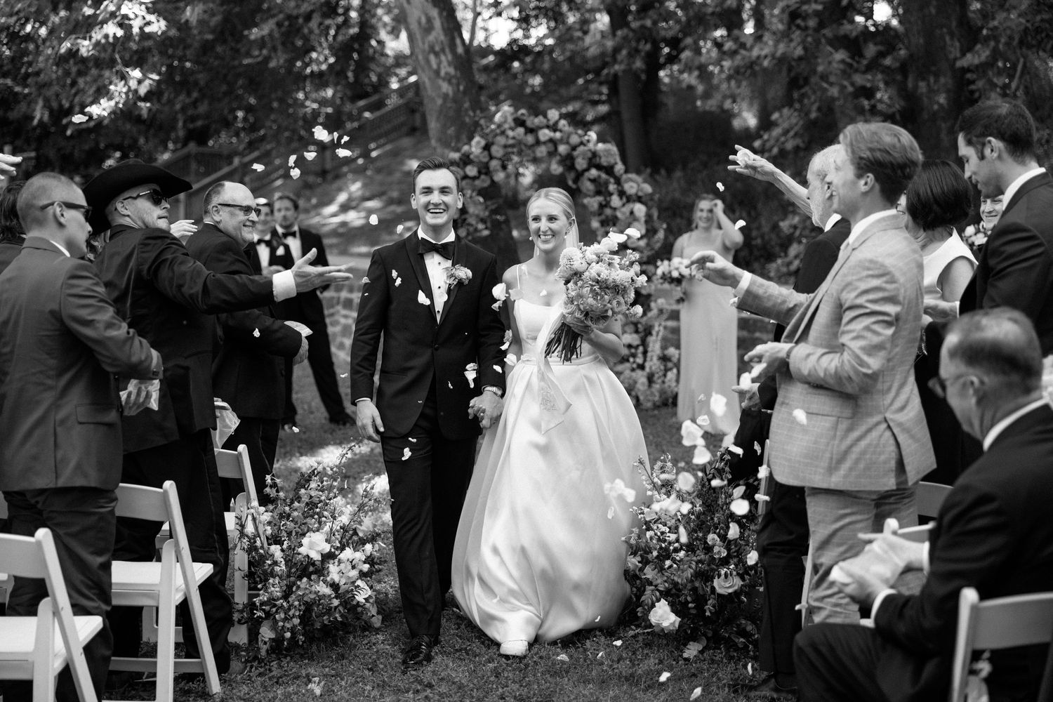 Black and white wedding ceremony scene with guests gathered outdoors.