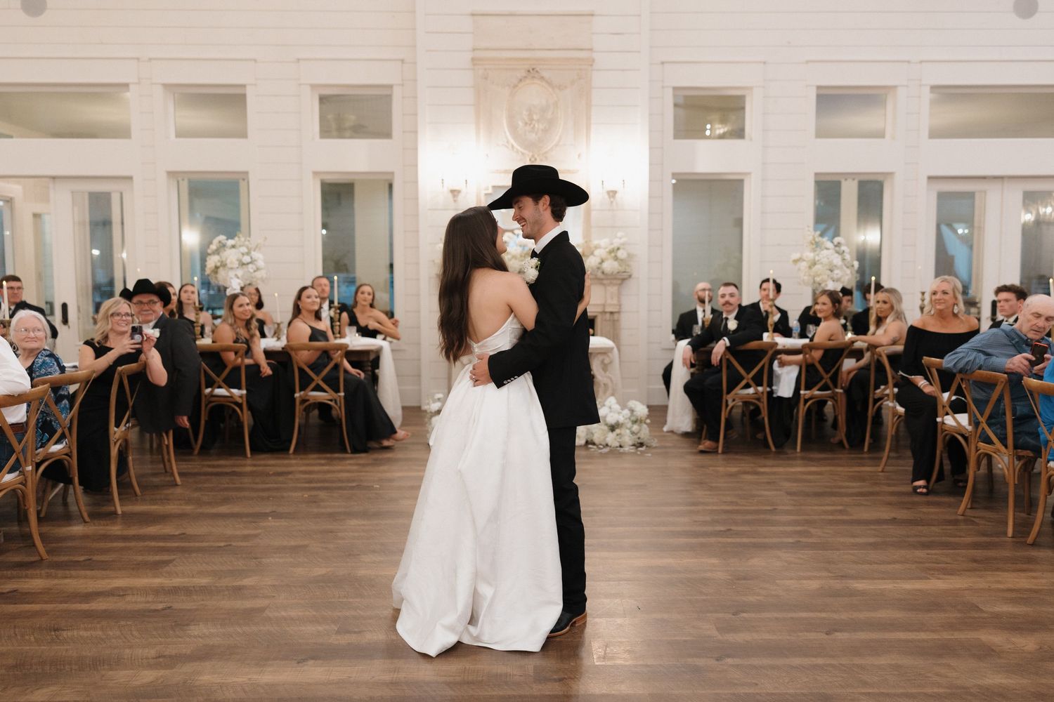 A wedding couple shares their first dance in a rustic venue with wooden floors and white walls as guests look on.