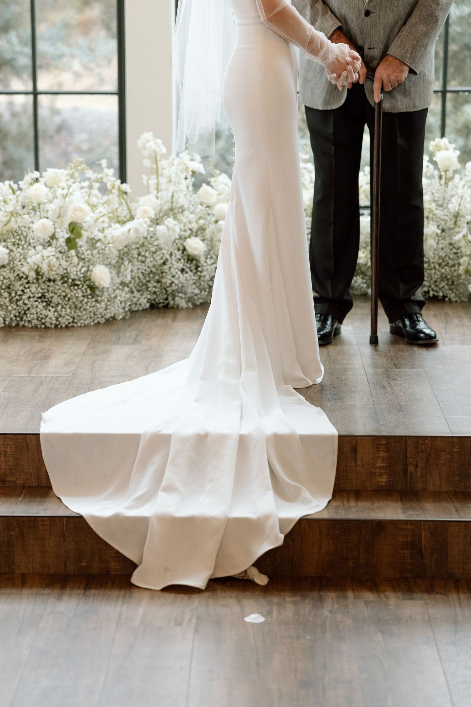 A flowing white wedding dress train cascades down wooden steps beside elegant black dress shoes at an indoor ceremony.