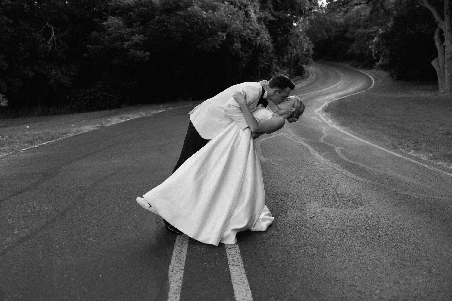 A romantic black and white photo of a couple embracing in a dip pose on a winding road surrounded by trees.