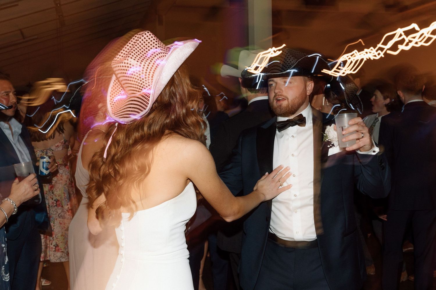 Wedding guests dancing together in formal attire on a dimly lit dance floor with string lights overhead.