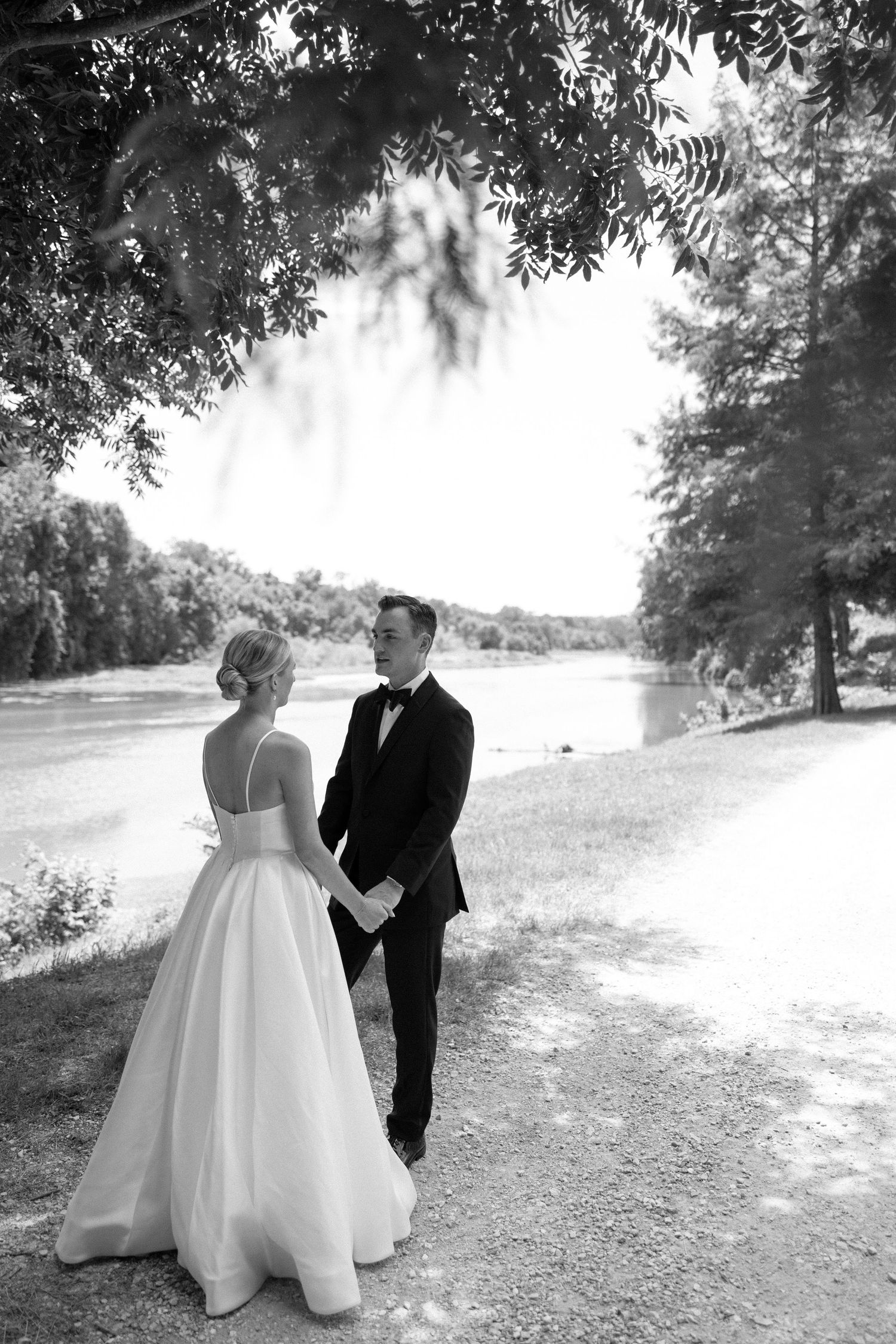 A couple in wedding attire stands together on a path by a serene lake under overhanging tree branches.