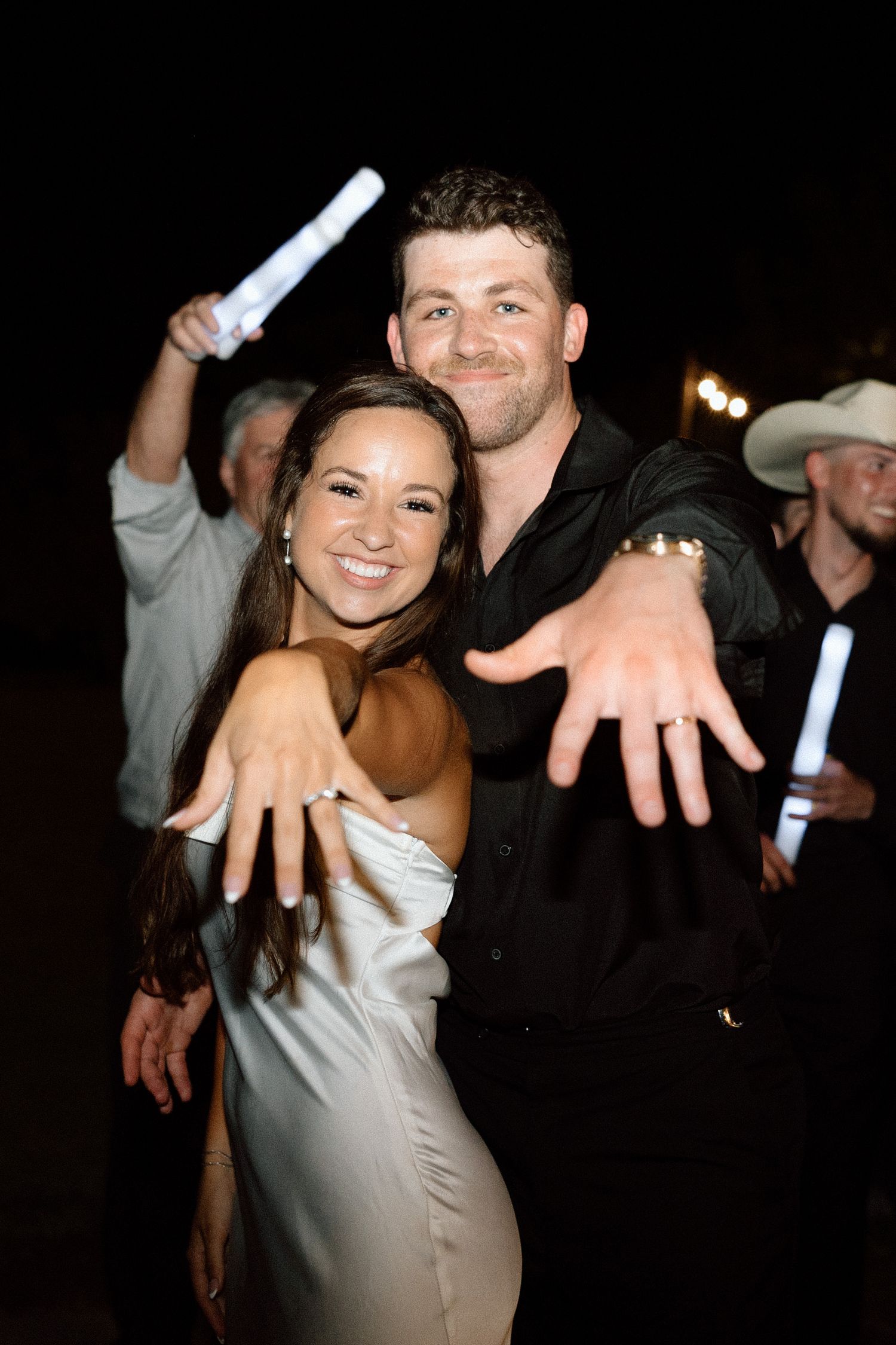 A couple in formal attire poses playfully at a wedding reception showing off their rings.