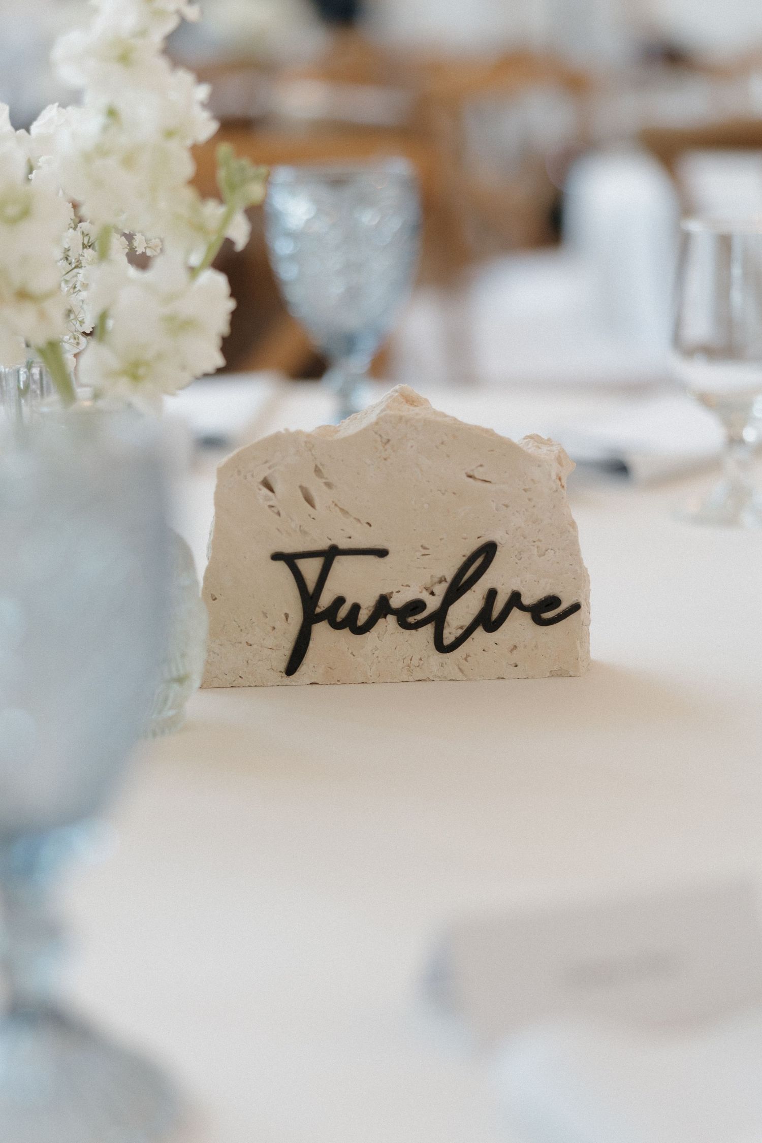 A rustic stone table number card displaying 'Twelve' with elegant calligraphy sits among blue glassware and white flowers.