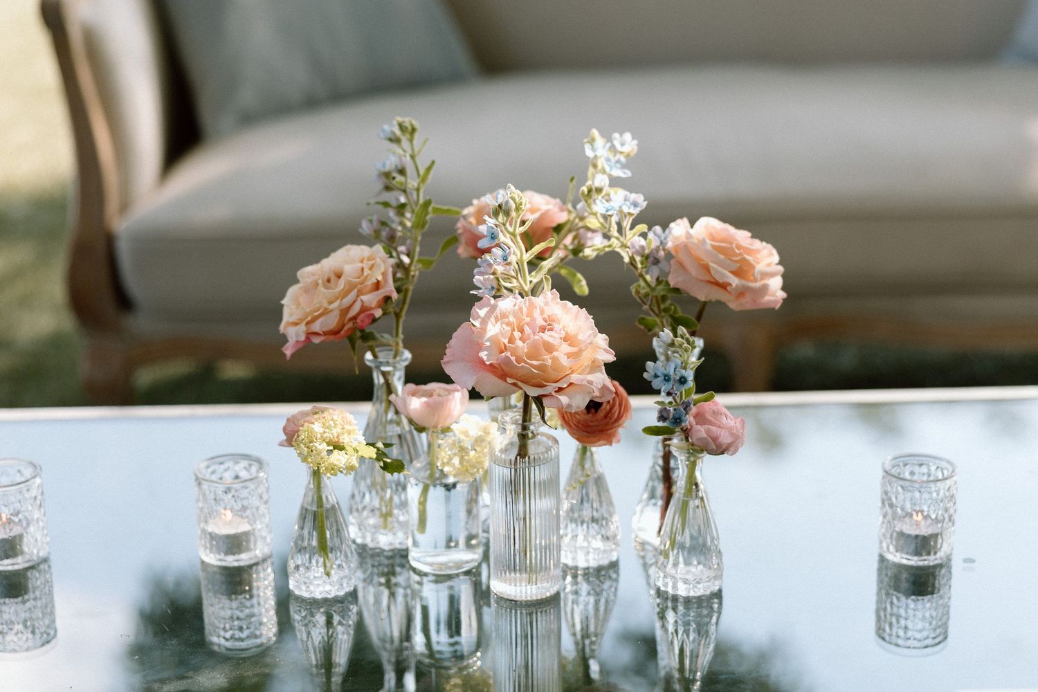 Glass vases filled with delicate pink and white flowers line up on a rustic wooden table for an elegant table setting.