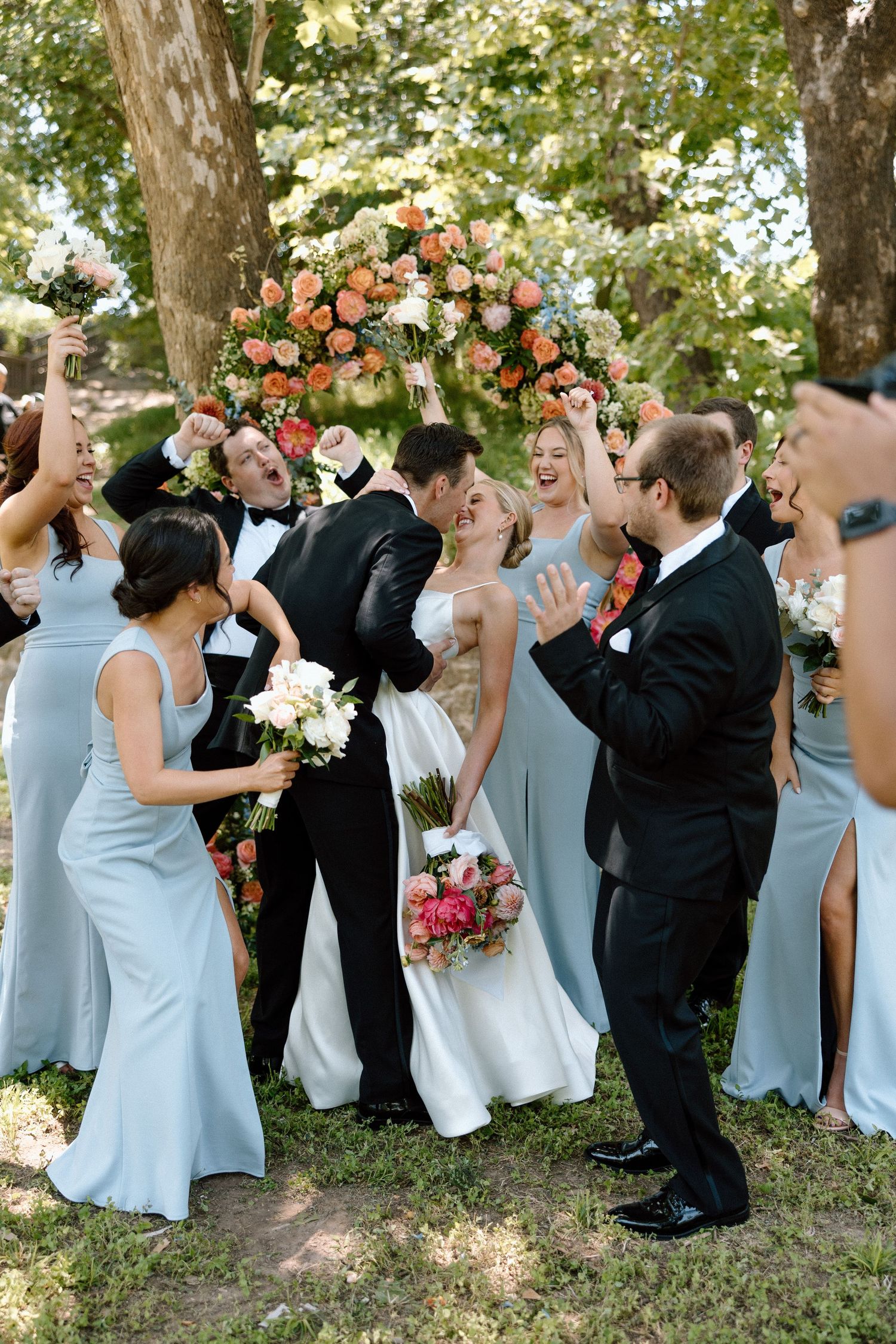 Wedding party celebrates under a beautiful floral arch with coral and white roses during an outdoor garden ceremony.