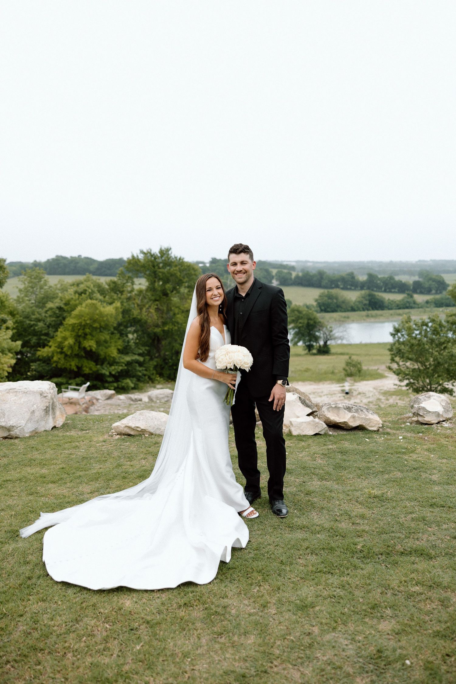 A bride and groom pose for a wedding photo on a grassy hilltop overlooking a scenic lake and rolling landscape.
