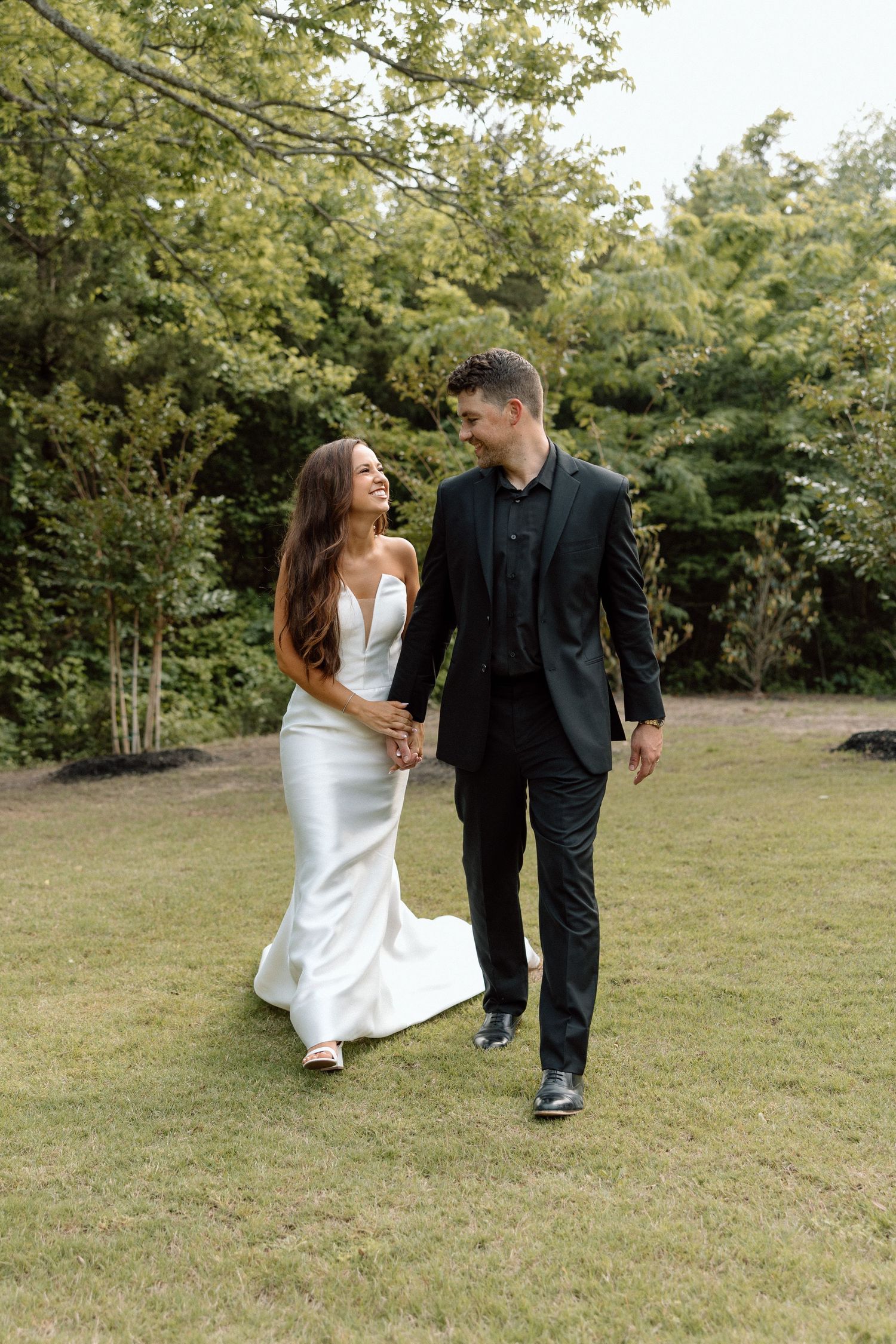 A couple in wedding attire walk together playfully across a grassy field on their special day.