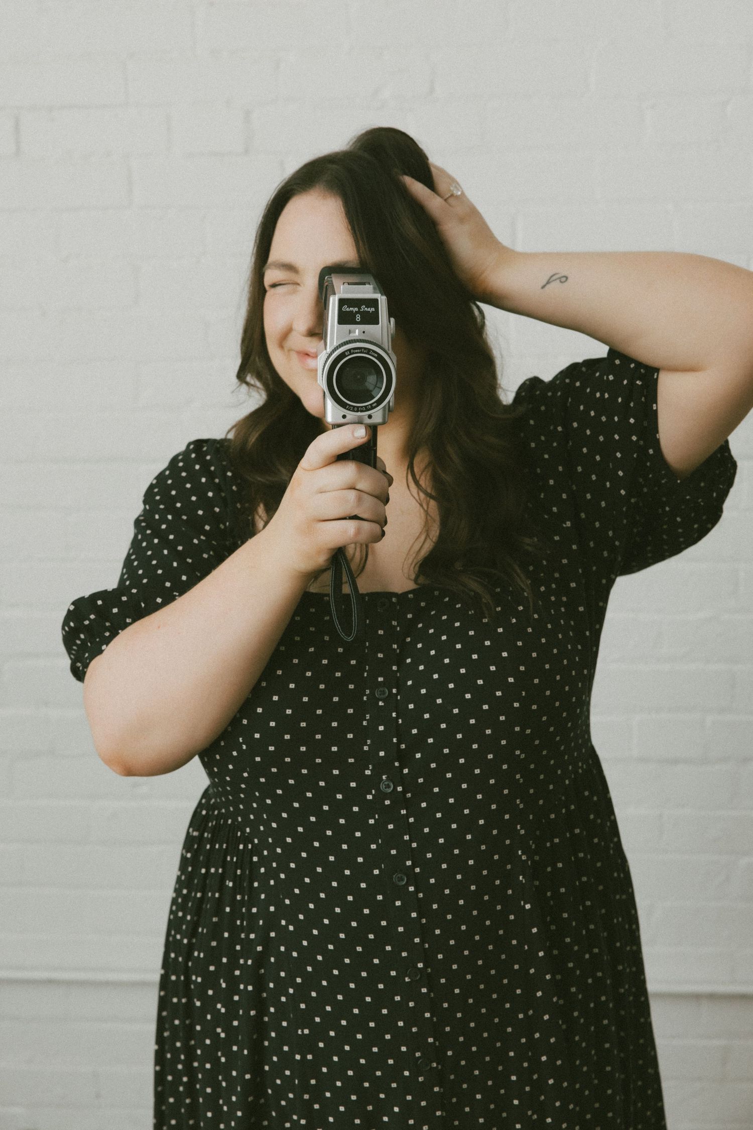 A person in a polka dot dress holds a vintage camera while posing against a white wall.