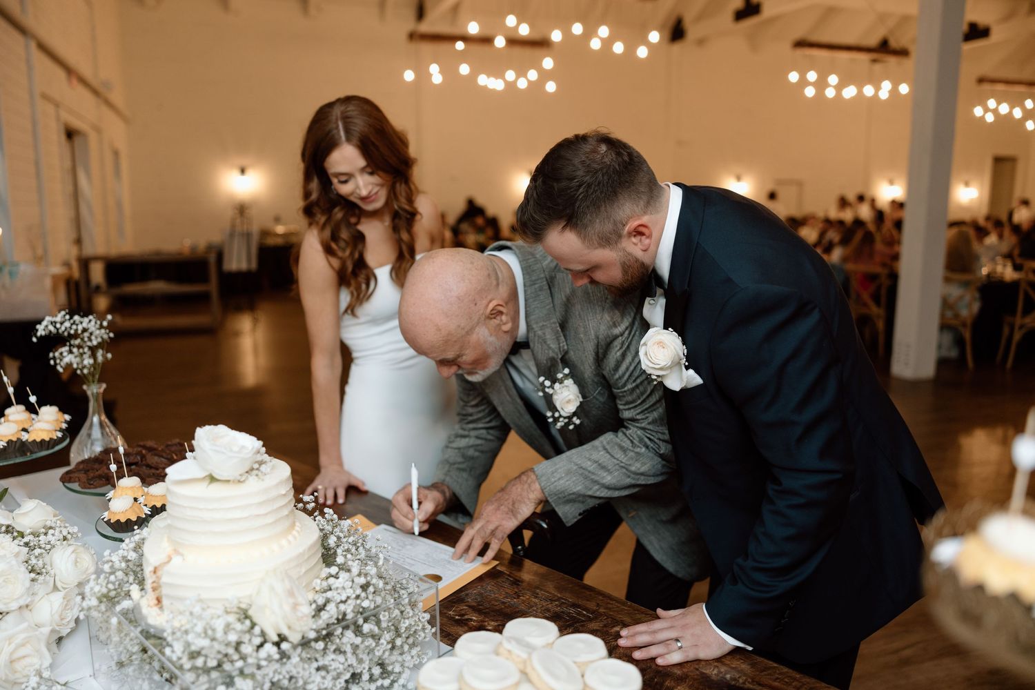 Three people cut into an elegant white wedding cake decorated with baby's breath flowers at an indoor reception.