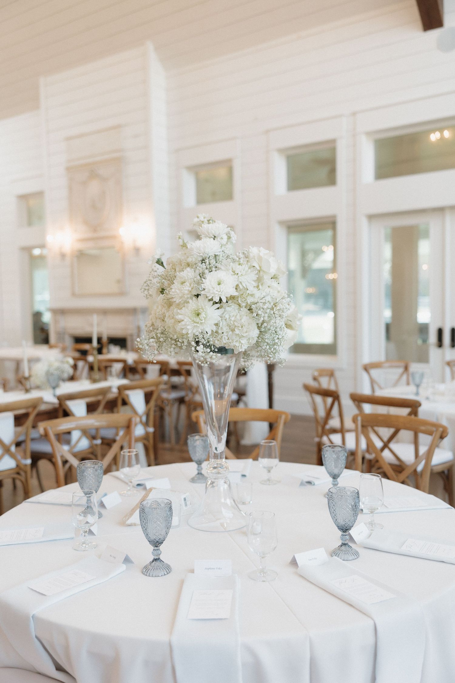 Elegant wedding reception venue with white round tables, cross-back chairs, and delicate white floral centerpieces.