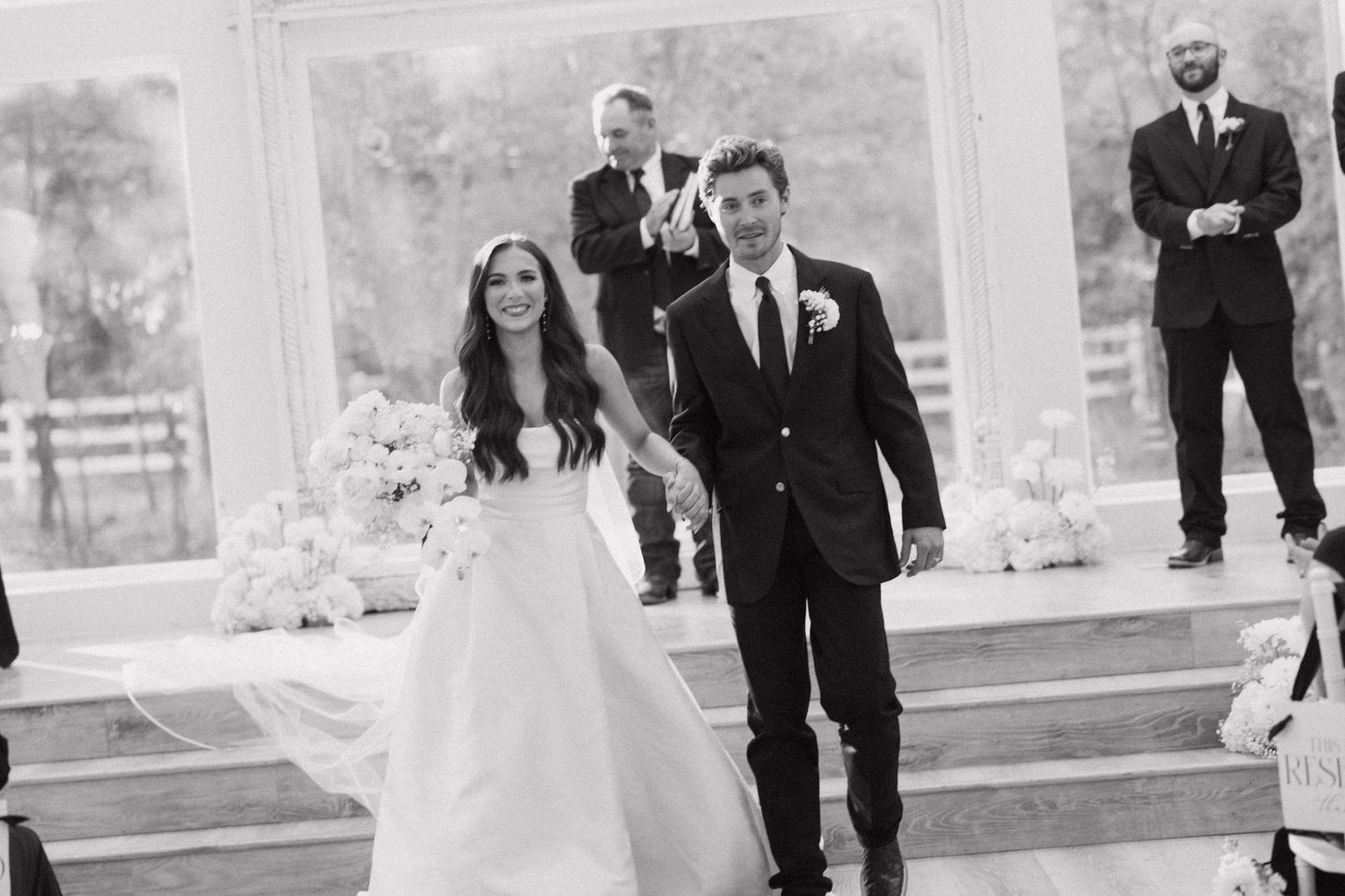 Black and white wedding party photo with people running and laughing outdoors.