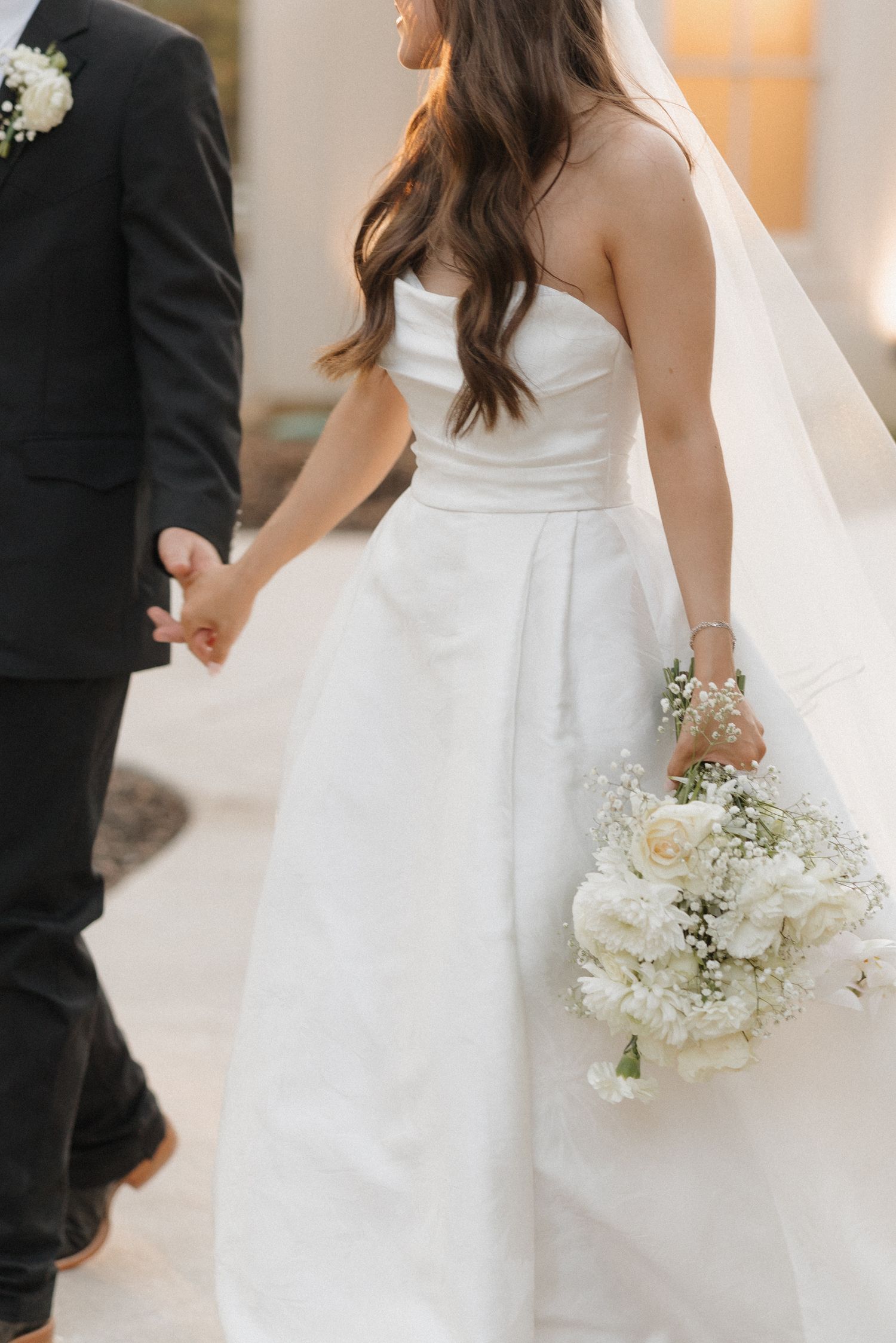 A bride in a white wedding dress holds a delicate bouquet of white roses and baby's breath at her side.