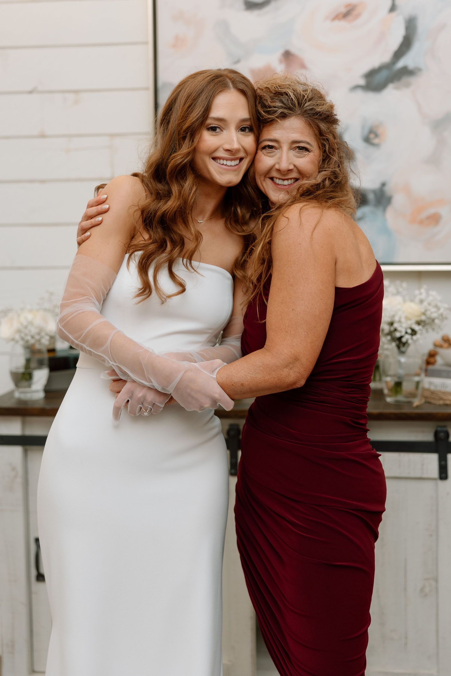 Two people embrace warmly in formal attire, one wearing a white dress and the other in a burgundy gown.