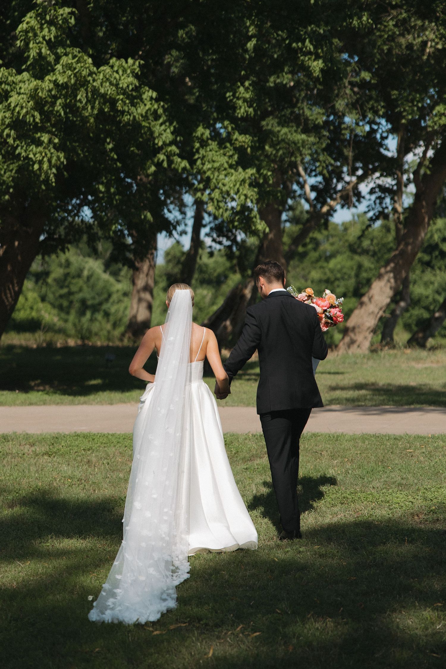 A bride and groom walk hand in hand through a lush green park on their wedding day.