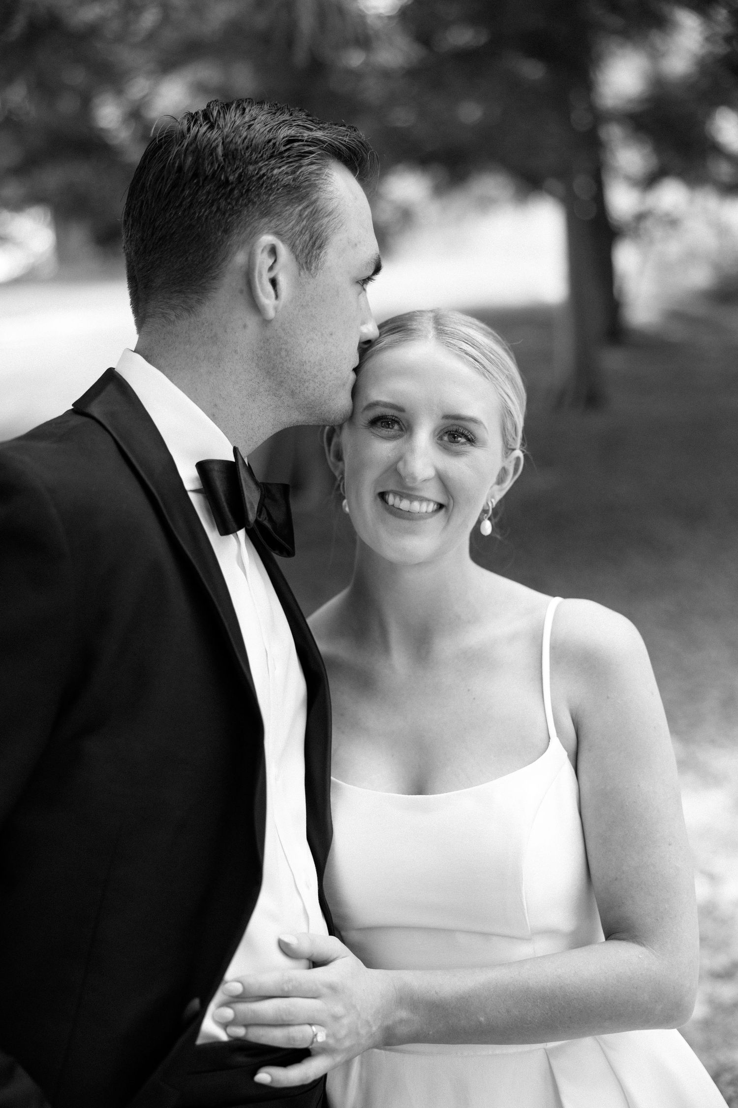 A black and white wedding portrait showing a couple embracing outdoors with trees in the background.