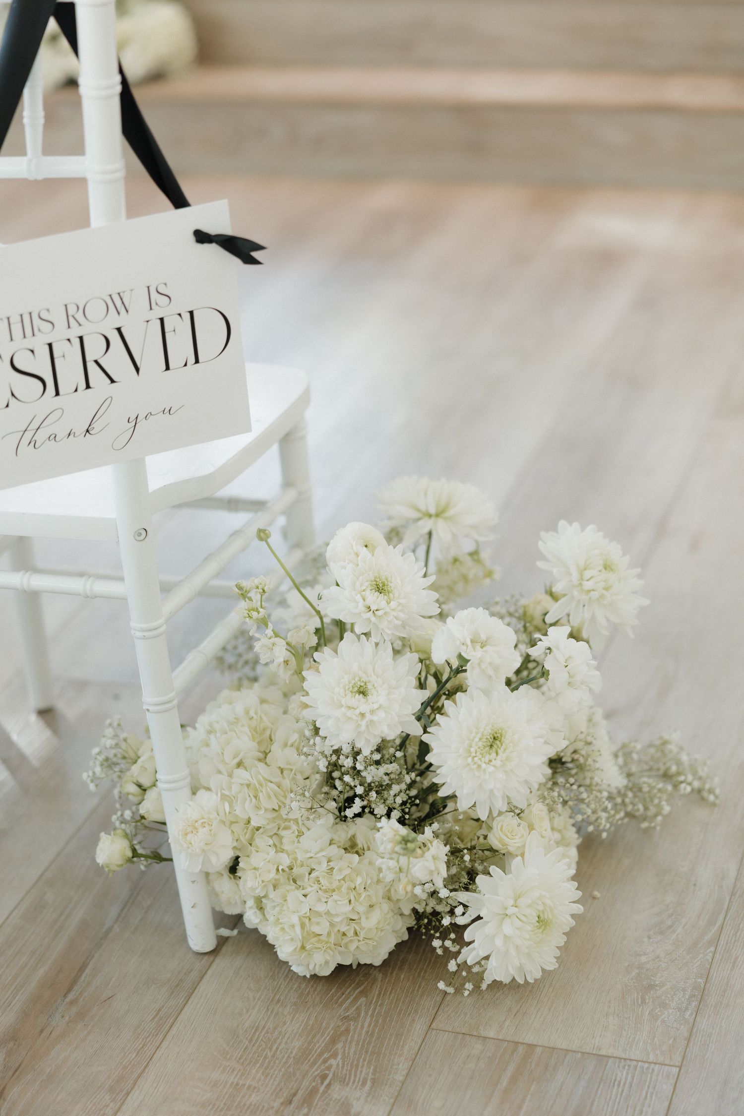 White floral arrangement with chrysanthemums and baby's breath sits beside a chair with a 'Reserved' sign on wooden flooring.